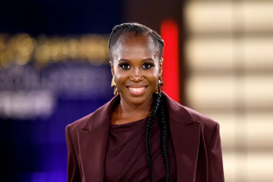 Motsi Mabuse Motsi Mabuse lächelt in die Kamera. Sie trägt einen weinroten Blazer, goldene Ohrringe und lange geflochtene Zöpfe.