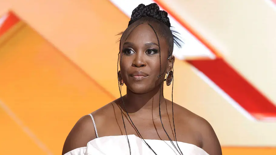 Motsi Mabuse Bruder Motsi Mabuse trägt ein weißes, schulterfreies Kleid mit voluminösen Ärmeln und einen geflochtenen Dutt vor orangefarbener Kulisse.
