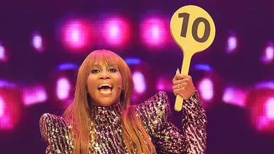 In einem glitzernden Kleid hebt Motsi Mabuse die 10-Punkte-Kelle von „Let’s Dance“ in die Luft und scheint gerade ihre Bewertung anzusagen. - Foto: IMAGO / Bildagentur Monn