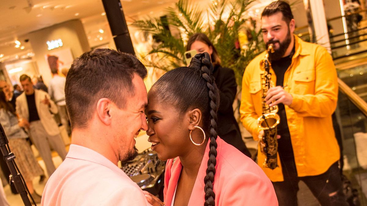 Motsi Mabuse: Meilenstein mit Evgenij Voznyuk Motsi Mabuse kuschet sich verliebt mit ihrem Ehemann Evgenij Voznyuk. Die beiden tragen zusammenpassende Outfits, im Hintergrund spielt ein Mann Saxofon.