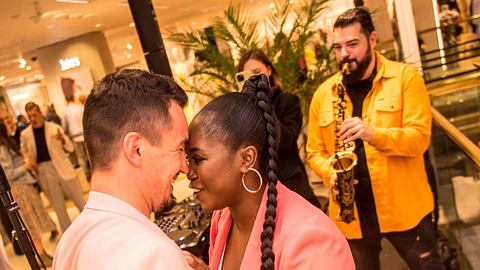 Motsi Mabuse kuschet sich verliebt mit ihrem Ehemann Evgenij Voznyuk. Die beiden tragen zusammenpassende Outfits, im Hintergrund spielt ein Mann Saxofon.  - Foto:  IMAGO / Hartenfelser