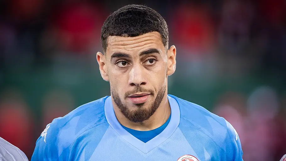 Ein Fußballspieler der tunesischen Nationalmannschaft trägt ein hellblaues Torwarttrikot und blickt vor dem Anpfiff konzentriert nach vorn. Der Hintergrund des Stadions ist unscharf.