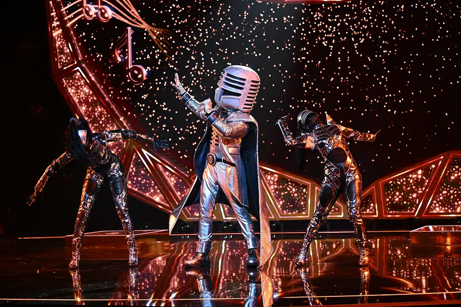 Mr. Mic ist ein lebendig gewordenes Mikrofon und verbindet seinen futuristischen Bühnen-Look mit einer großen Leidenschaft für Musik. Mr. Mic steht mit Mikrofonhelm und silbernem Kostüm zwischen zwei Tanzenden auf einer mit Musiknoten dekorierten Bühne.