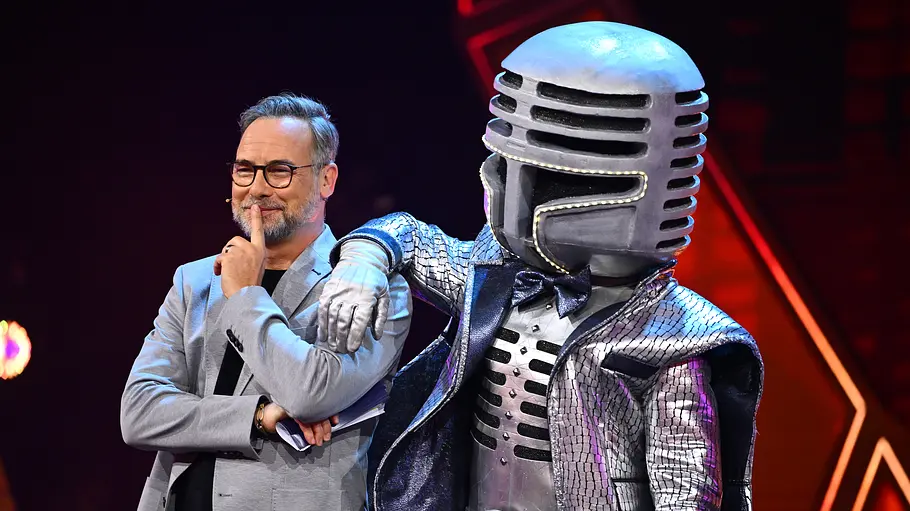Mr. Mic Ronan Keating Masked Singer Matthias Opdenhövel steht auf einer Bühne neben Mr. Mic im silbernen Mikrofonkostüm, der einen Arm auf seine Schulter legt.
