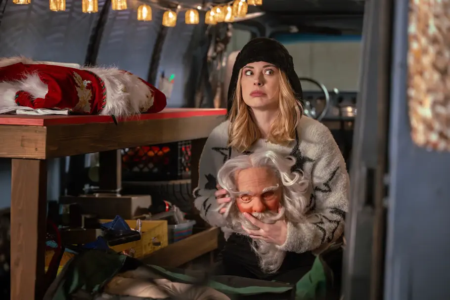 My Secret Santa Netflix Alexandra Breckenridge als Taylor hat eine Weihnachtsmann-Maske in der Hand. Sie ist in einem Lagerraum. Sie schaut, als wäre sie erwischt worden.