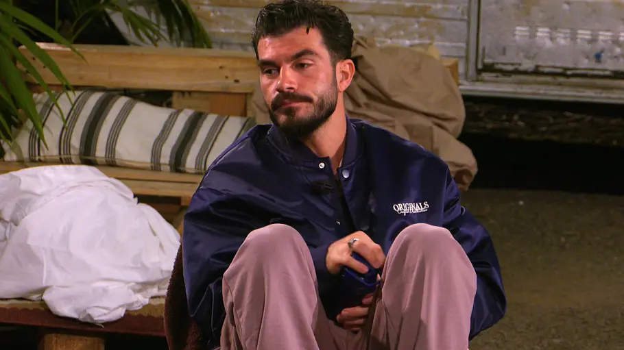 Maki Maki sitzt nachdenklich auf einem Bett in einem schlicht eingerichteten Raum, trägt eine dunkelblaue Jacke und helle Hose und blickt mit ernstem Gesichtsausdruck zur Seite.
