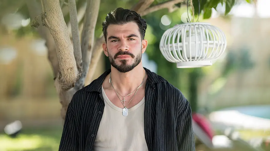 Maki Porträt von Maki, der unter einem Baum in die Kamera blickt. Er trägt ein schwarzes Hemd über einem hellen Shirt und mehrere Ketten, im Hintergrund sind ein Garten und eine weiße Laterne zu sehen.