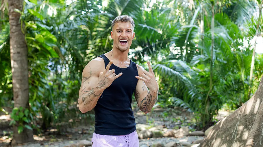 Laurenz Pesch Laurenz steht lachend im Dschungel vor tropischen Pflanzen. Er trägt ein schwarzes Tanktop und lilafarbene Shorts und zeigt mit beiden Händen das Rockerzeichen. Die Stimmung ist locker und entspannt.