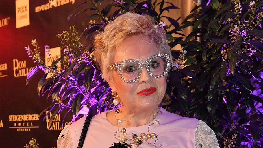 Maritta Emser Ältere Frau mit kurzen blonden Haaren und auffälliger, glitzernder Brille posiert auf einem Event vor einer beleuchteten Sponsorenwand, trägt ein rosafarbenes Kleid mit schwarzer Blumenapplikation, Statement-Kette und schwarze Handtasche.