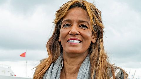 Nadja Abd el Farrag lächelt in die Kamera. Sie steht am Hamburger Hafen, hinter ihr sind graue Wolken und eine rote Fahne zu sehen. - Foto: IMAGO / Chris Emil Janßen