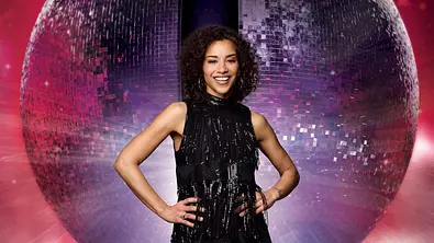 Eine Frau in einem schwarzen, fransigen Glitzerkleid und High Heels steht selbstbewusst vor einer großen, leuchtenden Discokugel auf rotem Hintergrund. - Foto: RTL / Jörn Strojny Eine Frau in einem schwarzen, fransigen Glitzerkleid und High Heels steht selbstbewusst vor einer großen, leuchtenden Discokugel auf rotem Hintergrund. - Foto: RTL / Jörn Strojny