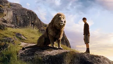 Szene aus Narnia: Ein Löwe sitzt neben einem Jungen auf einem Felsen in einer grasbewachsenen Landschaft unter wolkigem Himmel. - Foto: IMAGO / Allstar