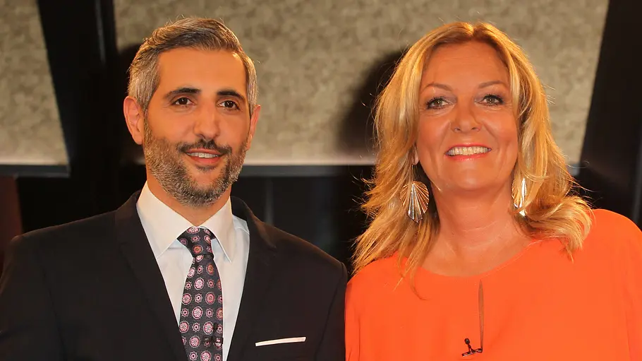 Bettina Tietjen und Michel Abdollahi Michel Abdollahi im dunklen Anzug und Bettina Tietjen im orangefarbenen Oberteil stehen lächelnd nebeneinander.