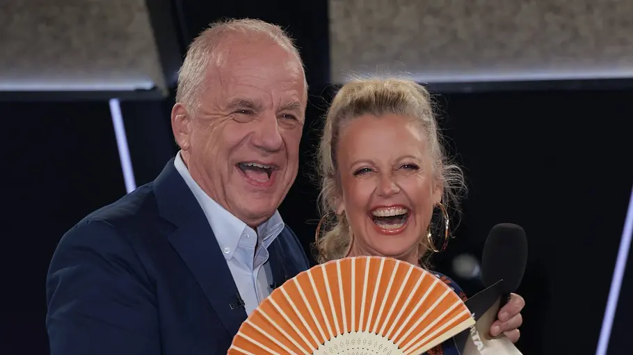 NDR Talkshow Barbara Schöneberger und Hubertus Meyer-Burckhardt stehen lachend auf der Bühne der „NDR Talk Show“. Hubertus trägt einen dunklen Anzug und hält einen orangefarbenen Fächer, Barbara trägt ein gemustertes Kleid; im Hintergrund sind Publikum und Studiolichter zu sehen.