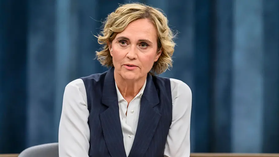 Caren Miosga Caren Miosga sitzt in weißer Bluse und dunkelblauer Weste an einem Tisch mit Papieren und Glas vor blauem Hintergrund.