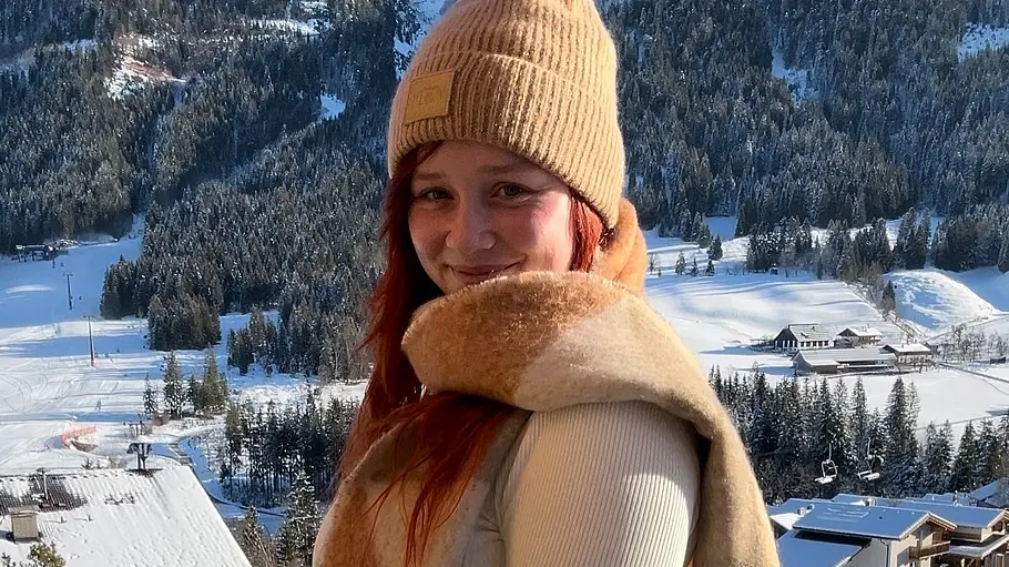 Cindy-Adriana Speich steht mit Strickmütze und großem Schal vor einer verschneiten Berglandschaft mit Häusern und Nadelwald.