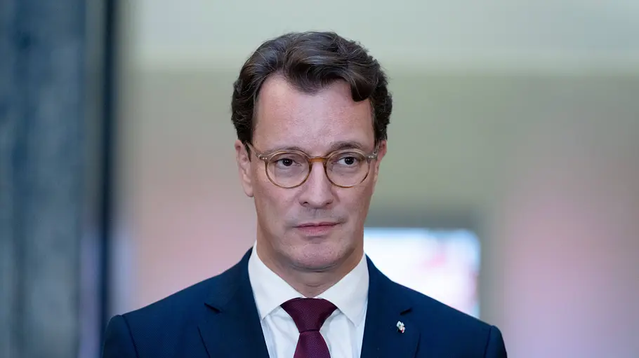 Hendrik Wüst Hendrik Wüst trägt eine runde Brille, einen dunkelblauen Anzug, ein weißes Hemd und eine bordeauxrote Krawatte.