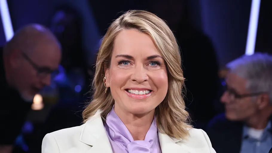 Jessy Wellmer lächelt in weißem Blazer und fliederfarbener Schluppenbluse. Hinter ihr sitzen unscharf weitere Personen.