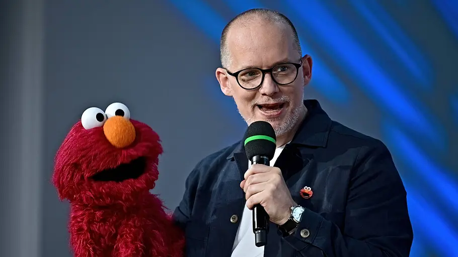 Martin Reinl Martin Reinl hält ein Mikrofon und eine rote Elmo-Handpuppe vor einem blau beleuchteten Hintergrund.