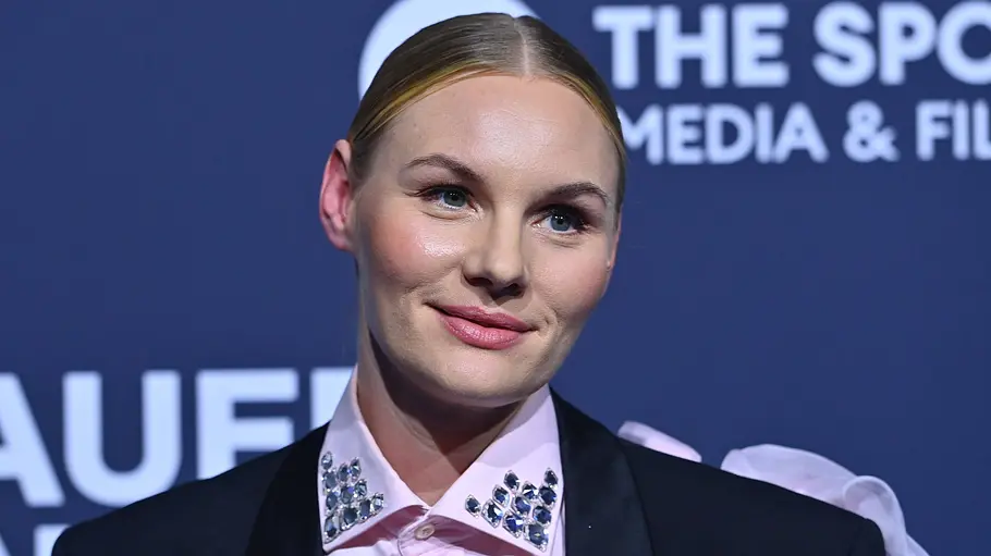 Rosalie Thomass Rosalie Thomass trägt einen schwarzen Blazer und ein rosa Hemd mit Schmucksteinen am Kragen vor einer blauen Schriftwand.