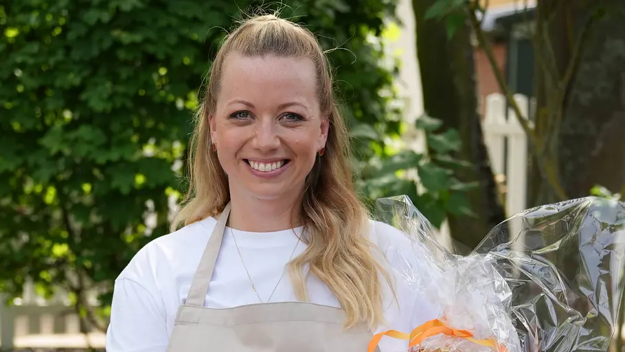 Zora Klipp Zora Klipp lächelt im Freien in einer beigen Schürze und hält ein in Folie verpacktes Geschenk mit orangefarbenen Bändern.