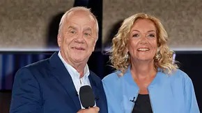 Zwei ältere Personen stehen nebeneinander in einem Fernsehstudio und blicken in die Kamera. Der Mann trägt ein dunkelblaues Sakko und hält ein Mikrofon in der Hand, die Frau trägt eine hellblaue Jacke über einem dunklen Oberteil. Beide lächeln, während der unscharfe Hintergrund auf eine TV- oder Bühnenproduktion hindeutet. - Foto: IMAGO / Future Image