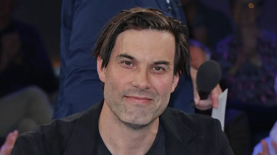 Max Simonischek sitzt im NDR-Studio in einem Sessel, trägt ein schwarzes Outfit und blickt ruhig in die Kamera.