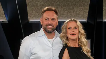 Barbara Schöneberger und Steven Gätjen stehen nebeneinander und lächeln in die Kamera; sie trägt ein schwarzes Kleid, er ein helles Hemd, im Hintergrund ist eine dunkle Studio-Kulisse. - Foto: IMAGO / BREUEL-BILD
