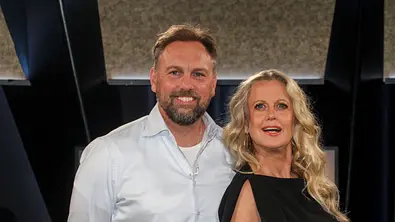 Barbara Schöneberger und Steven Gätjen stehen nebeneinander und lächeln in die Kamera; sie trägt ein schwarzes Kleid, er ein helles Hemd, im Hintergrund ist eine dunkle Studio-Kulisse. - Foto: IMAGO / BREUEL-BILD