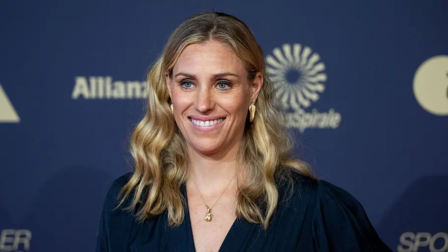 Angelique Kerber Eine Frau mit langen, welligen blonden Haaren lächelt vor einer Fotowand in die Kamera. Sie trägt ein dunkles Kleid sowie goldenen Schmuck und steht bei einer Veranstaltung, im Hintergrund sind Logos und Schriftzüge zu sehen.