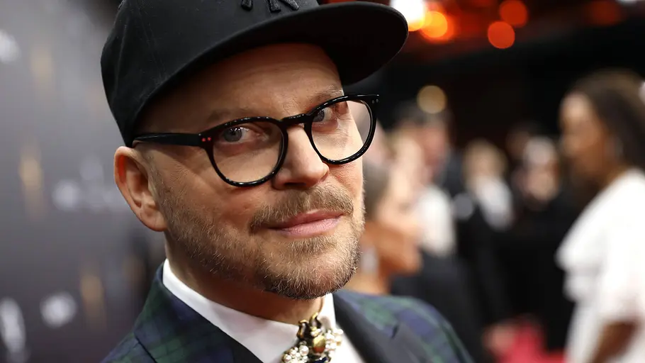 Armin Morbach Ein Mann mit Brille und schwarzer Kappe blickt leicht zur Kamera und lächelt dezent. Er trägt ein elegantes, kariertes Sakko mit Fliege und steht auf einem roten Teppich, während im Hintergrund unscharf weitere Gäste und Lichter einer Veranstaltung zu sehen sind.