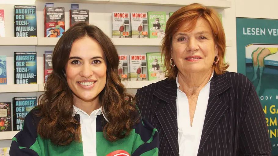 Christine Westermann und Mona Ameziane stehen lächelnd vor Bücherregalen, eine im Nadelstreifenblazer, die andere im Streifenpullover.