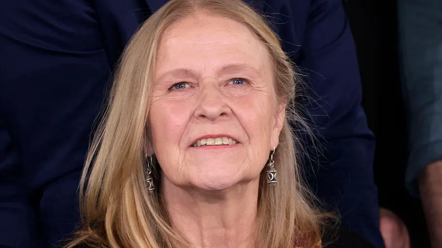 Cornelia Funke Eine ältere Frau mit schulterlangem, hellem Haar sitzt und lächelt freundlich in die Kamera. Sie trägt ein schwarzes Oberteil über einem gemusterten Kleid in Rot- und Goldtönen sowie Ohrringe. Die Aufnahme wirkt wie aus einer Talkshow oder Studioumgebung, im Hintergrund sind weitere Personen unscharf zu erkennen.