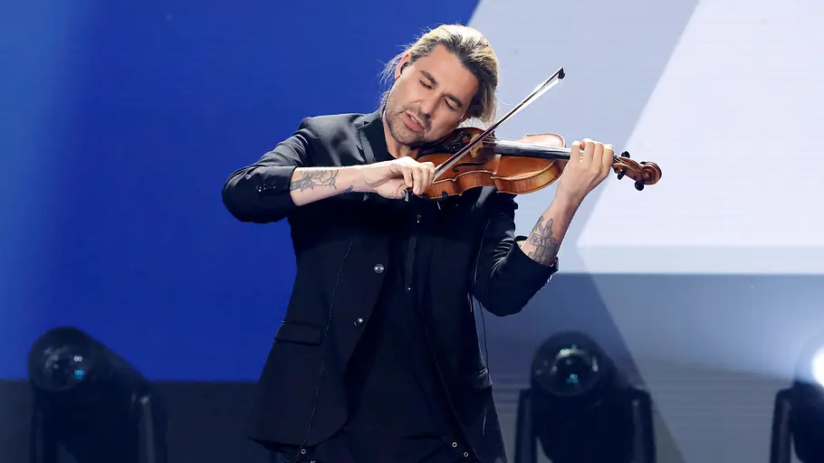 David Garrett Musiker mit langen, zurückgebundenen Haaren spielt konzentriert eine Geige auf einer Bühne. Er trägt ein schwarzes Outfit mit Sakko, während helle Scheinwerfer und Nebeleffekte die Szene beleuchten. Im Hintergrund sorgen blau-weiße Lichtelemente für eine moderne Konzertatmosphäre.
