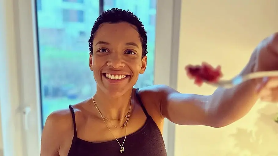 Dr. Shiwa Soukou Lächelnde Person mit kurzem, lockigem Haar steht in einer hellen Küche oder Essbereich vor einer großen Glastür. Sie trägt ein schwarzes Trägertop und hält ein Stück rotes Obst in der Hand, während sie ein weiteres in Richtung Kamera reicht. Im Hintergrund sind Gläser, eine Arbeitsfläche und unscharfe Wohngebäude außerhalb des Fensters zu sehen.