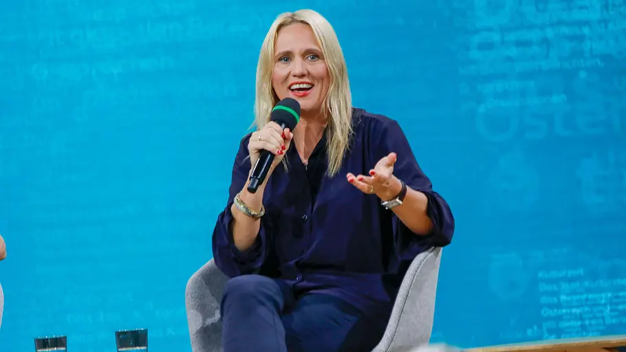 Elisa Hoven Blonde Frau sitzt auf einem Podium und spricht mit einem Mikrofon in der Hand. Sie trägt ein dunkelblaues Outfit und gestikuliert während ihrer Rede. Im Hintergrund ist eine blau beleuchtete Bühne zu sehen, vor ihr sitzen Zuhörerinnen und Zuhörer.