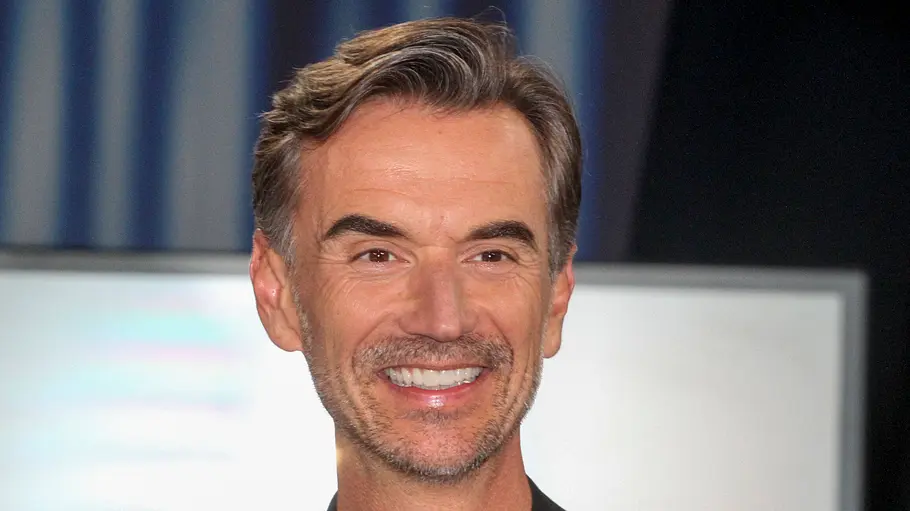 Florian Silbereisen Ein Mann mit graumeliertem Haar und leichtem Bart lächelt in die Kamera. Er trägt ein dunkles Sakko und befindet sich offenbar in einer Studio- oder Eventumgebung, im Hintergrund ist unscharf eine Leinwand oder ein Bildschirm zu erkennen.
