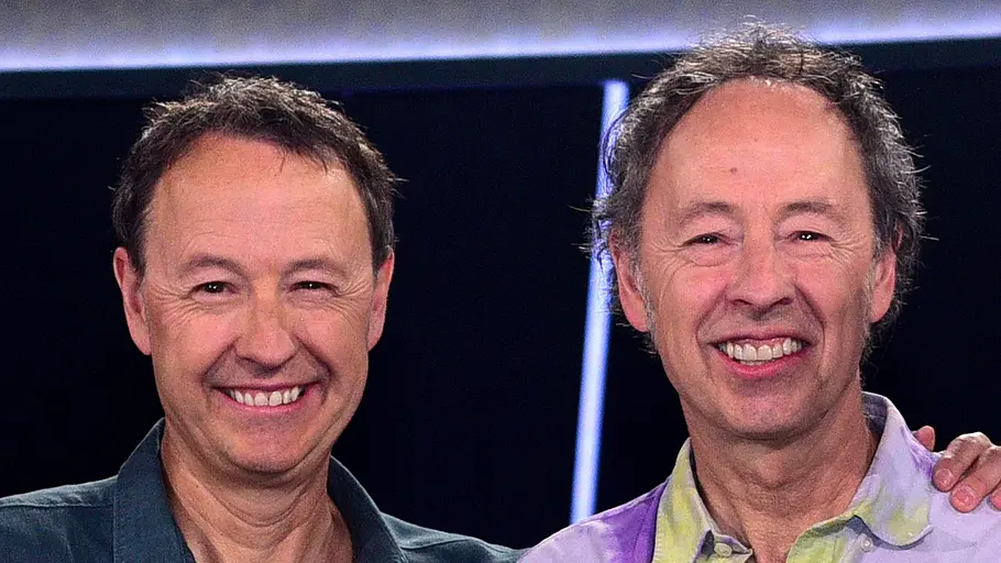 Gerrit und Frederik Braun Zwei Männer stehen nebeneinander und lächeln in die Kamera. Der eine trägt ein dunkles Hemd, der andere ein bunt gemustertes Shirt. Einer legt dem anderen freundschaftlich die Hand auf die Schulter. Die Szene wirkt wie aus einer TV- oder Studioaufnahme mit dunklem Hintergrund und Lichtakzenten.