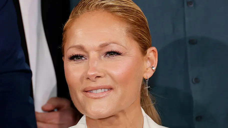 Helene Fischer Eine Frau mit zurückgebundenem, blondem Haar lächelt leicht in die Kamera. Sie trägt ein helles Blazer-Outfit und dezente Schmuckstücke, darunter Ohrringe. Die Aufnahme wirkt wie aus einem Studio oder einer TV-Produktion, im Hintergrund sind unscharf weitere Personen zu erkennen.