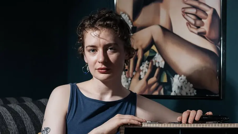 Katharina Raab Junge Person mit kurzen, lockigen Haaren sitzt in einem dunkel eingerichteten Raum neben einem detailreichen Miniaturmodell in einem geöffneten Koffer. Sie trägt ein ärmelloses dunkelblaues Oberteil und hat mehrere Tattoos an Armen und Beinen. Im Hintergrund hängt ein großformatiges Kunstposter mit zwei Frauenporträts an der Wand, während gedämpftes Licht für eine ruhige, kreative Atmosphäre sorgt.