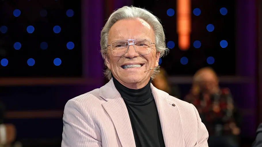 Wolfgang Lippert Älterer Mann mit grauem Haar und großer Brille sitzt in einer TV-Studio-Umgebung und lächelt. Er trägt einen hellrosa Blazer über einem schwarzen Rollkragenpullover. Im Hintergrund sind unscharfe Zuschauer sowie dekorative Lichtpunkte in Blau und Violett zu sehen.