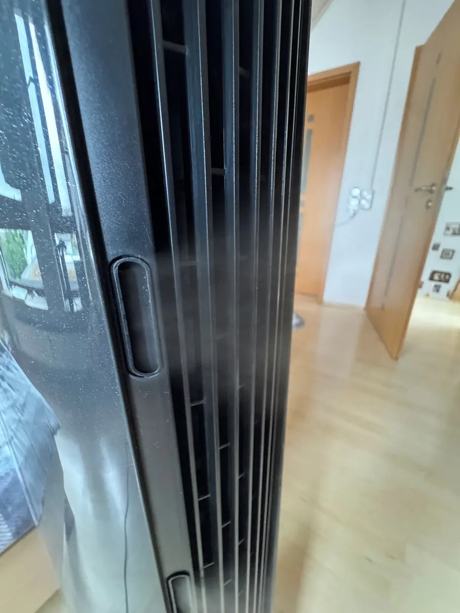 Nebel Nahaufnahme eines schwarzen Turmventilators, aus dessen schmalen Luftschlitzen feiner Nebel austritt. Im Hintergrund sind ein heller Holzfußboden und eine geöffnete Zimmertür zu sehen.