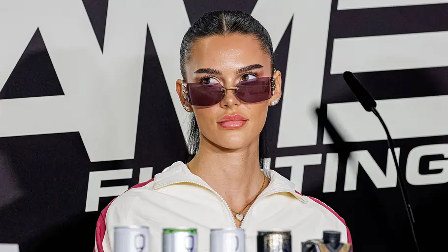 Nelly Noire Fame Fighting Nelly Noire mit Sonnenbrille und weißer Sportjacke sitzt hinter Getränkedosen. Neben ihr steht ein Mikrofon vor einer Logowand.