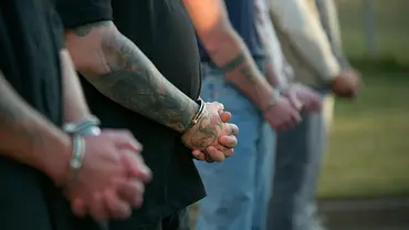 Mehrere Personen stehen mit Handschellen nebeneinander. Im Fokus sind tätowierte Arme und verschränkte Hände vor dem Körper. - Foto: © 2026 Netflix, Inc. All Rights Reserved. Mehrere Personen stehen mit Handschellen nebeneinander. Im Fokus sind tätowierte Arme und verschränkte Hände vor dem Körper. - Foto: © 2026 Netflix, Inc. All Rights Reserved.