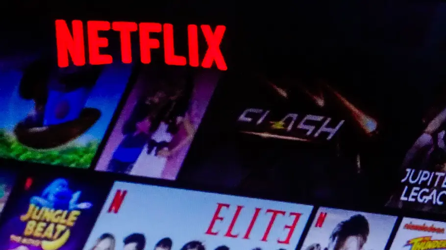 Netflix ändert Programm Auf dem Bild sind der rote Netflix-Schriftzug auf einem Smartphone sowie verschiedene Serien- und Filmsymbole auf einem Fernseher im Hintergrund zu sehen. Darunter sind Titel wie „Elite“, „Jupiter's Legacy“ und „Money Heist“. Rechts im Bild ist der Schriftzug „Unlimited movies, shows, and more“ zu lesen.