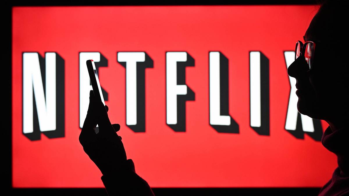 Netflix-House nicht in Deutschland Ein Mann mit Brille steht vor einem Bildschirm, auf dem das Netflix-Logo zu sehen ist und starrt auf sein Handy
