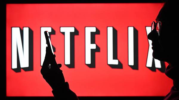 Ein Mann mit Brille steht vor einem Bildschirm, auf dem das Netflix-Logo zu sehen ist und starrt auf sein Handy - Foto:  IMAGO / NurPhoto