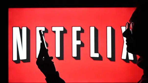Ein Mann mit Brille steht vor einem Bildschirm, auf dem das Netflix-Logo zu sehen ist und starrt auf sein Handy - Foto: IMAGO / NurPhoto Ein Mann mit Brille steht vor einem Bildschirm, auf dem das Netflix-Logo zu sehen ist und starrt auf sein Handy - Foto: IMAGO / NurPhoto