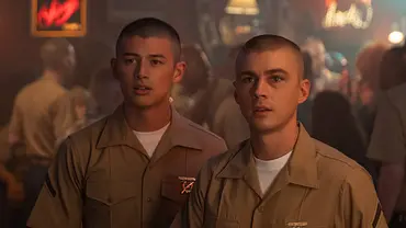Zwei junge Männer in khakifarbenen Marineuniformen stehen nebeneinander in einer Bar mit schummrigem Licht. Beide haben sehr kurze Haare und schauen aufmerksam nach vorne. Im Hintergrund sieht man weitere Menschen und verschwommene Lichter einer belebten Bar-Atmosphäre. - Foto: Patti Perret/Netflix © 2024 Zwei junge Männer in khakifarbenen Marineuniformen stehen nebeneinander in einer Bar mit schummrigem Licht. Beide haben sehr kurze Haare und schauen aufmerksam nach vorne. Im Hintergrund sieht man weitere Menschen und verschwommene Lichter einer belebten Bar-Atmosphäre. - Foto: Patti Perret/Netflix © 2024