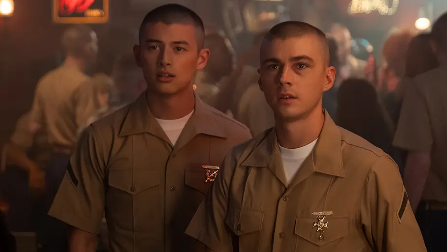 Netflix-Serie „Boots“ Zwei junge Männer in khakifarbenen Marineuniformen stehen nebeneinander in einer Bar mit schummrigem Licht. Beide haben sehr kurze Haare und schauen aufmerksam nach vorne. Im Hintergrund sieht man weitere Menschen und verschwommene Lichter einer belebten Bar-Atmosphäre.